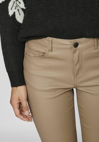 Bež hlače, prilegajoče se s gladko teksturo in standardnim dizajnom s petimi žepi. Roka počiva ob strani blizu pasu.