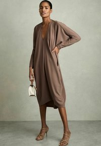 Brun, långärmad klänning med V-ringning, sidofickor och en flödande design. Matchad med nakna, strappy klackar och en liten vit handväska.