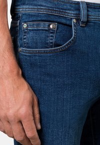 Dunkelblaue Denim-Jeans mit einer Nahaufnahme auf einer Vorderasche. Sie verfügen über Metallnieten und Kontrastnähte, die eine glatte Textur zeigen.