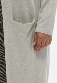 Γκρι ζακέτα από απαλό ύφασμα, με πλαϊνή τσέπη και μανσέτες με ρίγες. Ο σχεδιασμός είναι μακρύς και με χαλαρή εφαρμογή, παρουσιάζοντας μινιμαλιστική υφή.