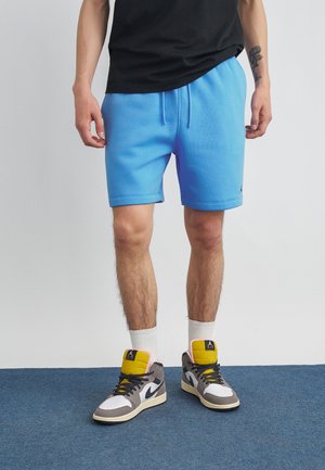 Isik kandmas eredat sinist lühikesi pükse, musta t-särki, valgeid sokke ja halli, valget, kollast ja musta Nike'i spordijalatseid, seisab sinisel vaibal.