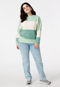 Pull en maille avec rayures vertes et crème, col rond, manches longues, associé à un jean denim bleu clair et des baskets grises.