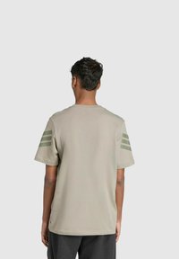Khaki T-Shirt mit kurzen Ärmeln und drei horizontalen olivgrünen Streifen auf der linken Schulter, aus weichem Baumwollstoff, lässiger Schnitt.