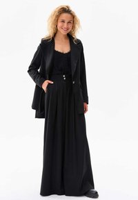 Zwarte op maat gemaakte blazer over een zwarte top met kantafwerking, gecombineerd met zwarte wijde broek met een knoopsluiting in de taille. Soepele stof, strakke ontwerp.