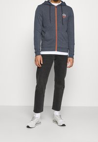 Marinblå zip-hoodie med en ljusorange dragkedja, två framfickor, justerbara snoddar och ett hexagonalt logomärke på bröstet. Svarta jeans och sneakers.