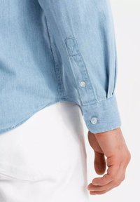 Chemise en denim bleu clair avec des poignets boutonnés, texture douce et ourlet arrondi, partiellement visible à côté d'une main sur un fond uni.