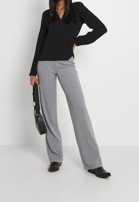 Černý top s dlouhým rukávem s výstřihem do V a nabíranými rameny, doplněný o šedé široké kalhoty. Modelka drží černou kabelku a má na sobě špičaté černé boty.