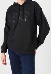 Felpa nera con cappuccio, dotata di tasca frontale e logo "GAP" con paillettes. Presenta una vestibilità comoda e polsini e orlo a costine. Tessuto morbido.