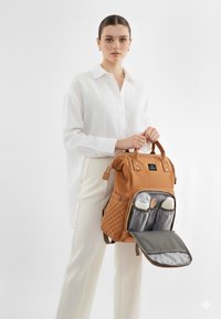 Tan rygsæk med teksturerede sider, dobbelt flaske lommer og lynlåslommer foran, holdt af en model i en hvid skjorte og cremefarvede bukser.