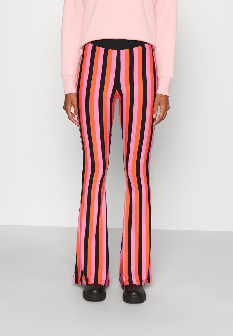 Personne portant un pantalon évasé taille haute à rayures verticales roses, orange, rouges et noires, associé à des chaussures noires et un haut rose clair.