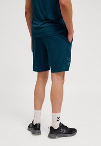 Bermudas atléticas de color verde azulado con un ajuste holgado, bolsillos laterales y un sutil detalle de logo. Llevadas con zapatos negros y calcetines blancos con franjas negras.