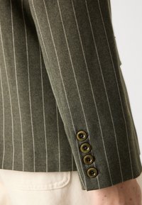 Oliivroheline triibuline bleiseri varrukas kontrastsete valgete peentriipudega. Varrukas on neli messingust nööpi, millel on tekstureeritud kangast viimistlus.