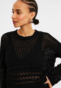 Schwarzer Strickpullover mit einem Lochmuster, strukturiertem Rippenkragen und aufwendigem Stichdesign über die Vorderseite und Ärmel.