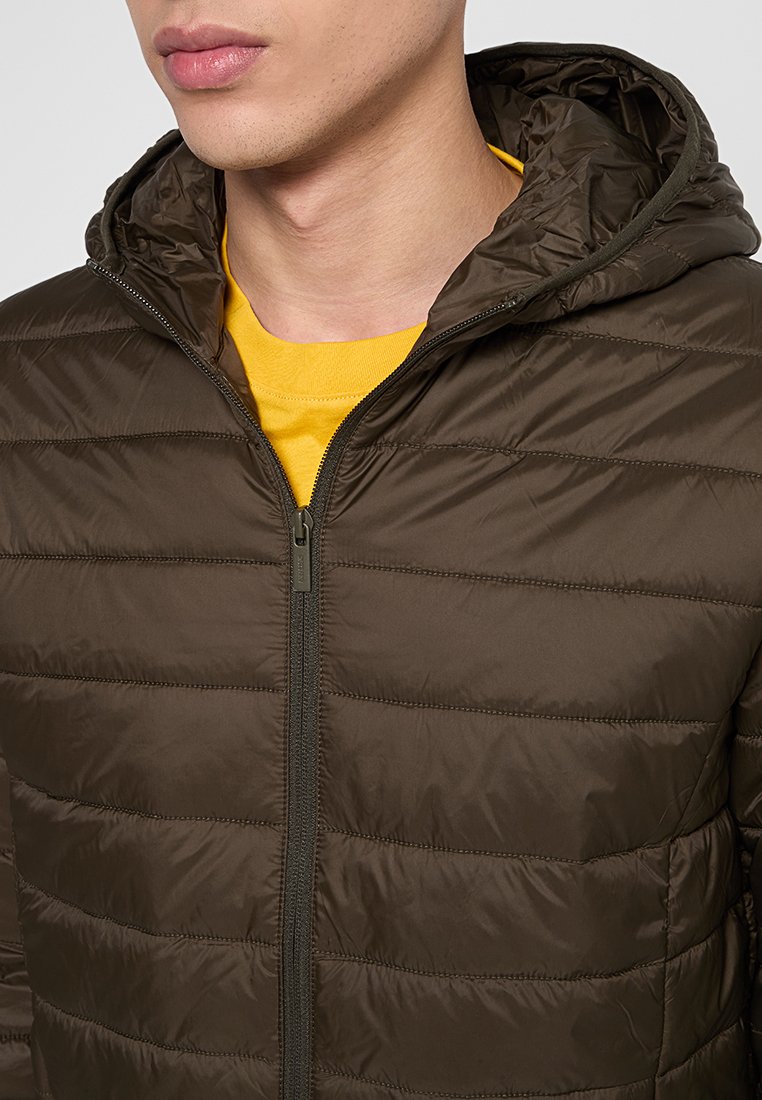 Veste matelassée vert foncé avec une capuche, présentant un motif matelassé horizontal et une fermeture éclair à l'avant. Portée sur une chemise jaune.