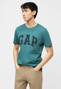 Νέος άνδρας με κοντά μαύρα μαλλιά, που φοράει μπλουζάκι GAP σε τιρκουάζ χρώμα και μπεζ παντελόνι, με τα χέρια στις τσέπες, κοιτάζει ελαφρώς προς τα αριστερά σε λευκό φόντο.