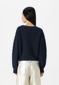Granatowy sweter z prążkowanej dzianiny z teksturowanymi, diamentowymi rękawami. Tył ma okrągły dekolt i luźny krój. Srebrne metaliczne spodnie.