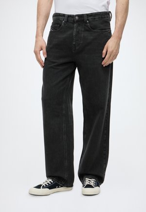 Personne portant un jean noir ample et des baskets noires basses avec des lacets et semelles blanches sur fond blanc.