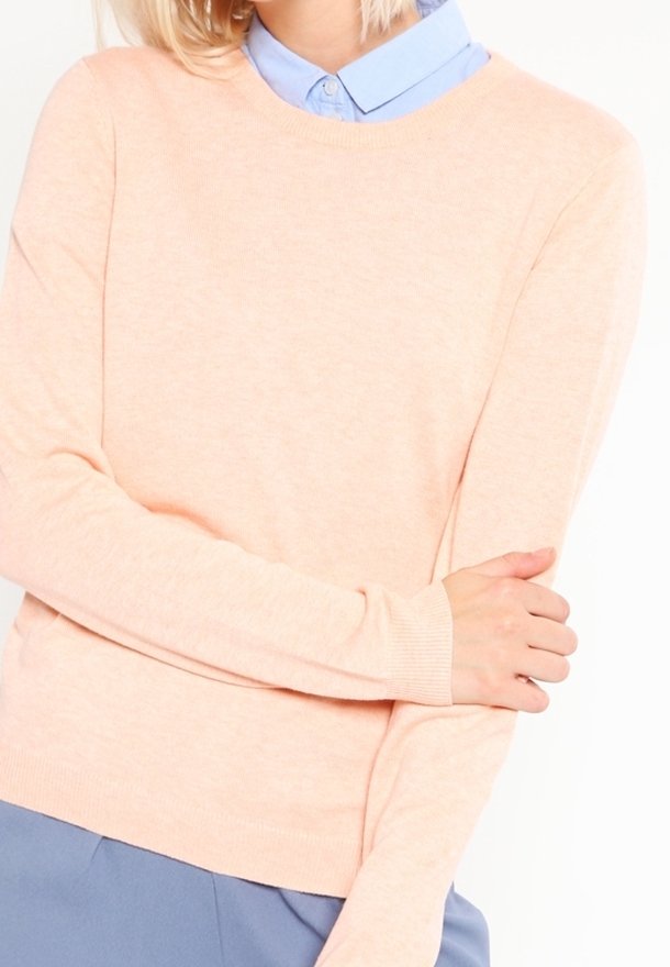 Pull en rose clair avec un col rond et des manches longues, fabriqué en tissu doux et texturé, superposé à une chemise à col bleu clair.