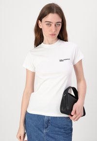 Witte T-shirt met korte mouwen en een ronde hals, met zwart "KARL LAGERFELD JEANS" logo op de borst. Gecombineerd met een spijkerrok en een zwarte tas.