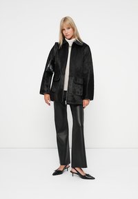 Giacca nera in pelliccia sintetica con chiusura a zip, forma oversize, due tasche frontali, indossata sopra un maglione chiaro, abbinata a pantaloni in pelle nera.