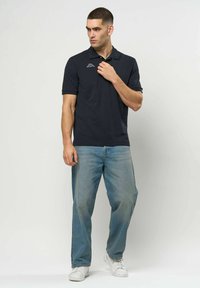 Mann i svart polotrøye, lyseblå jeans og hvite joggesko som står mot en ensfarget hvit bakgrunn og ser til siden.