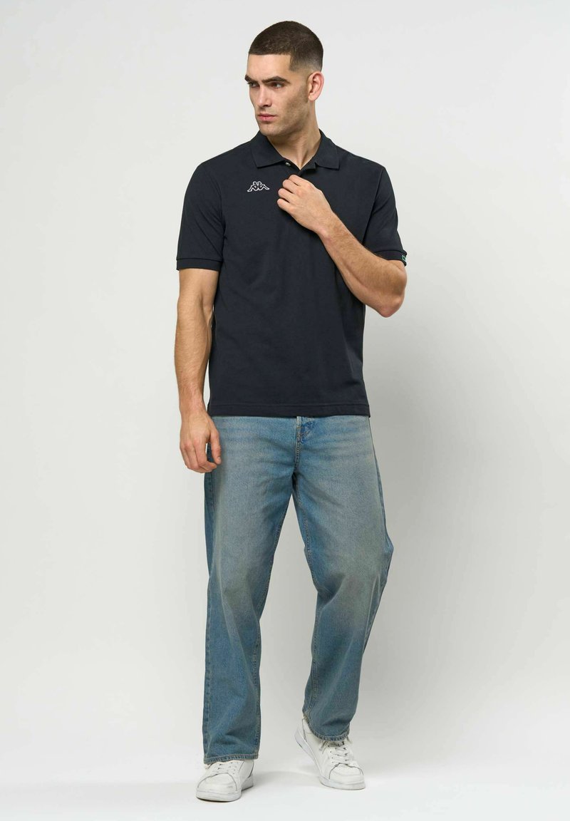 Mann i svart polotrøye, lyseblå jeans og hvite joggesko som står mot en ensfarget hvit bakgrunn og ser til siden.