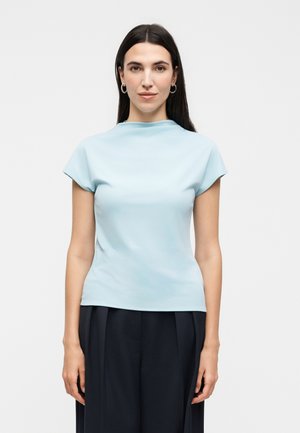 Jeune femme aux longs cheveux foncés, portant des boucles d’oreilles créoles, un haut bleu clair à manches courtes et un pantalon plissé foncé, debout devant un fond uni.