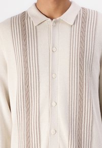 Light beige cardigan with a collared neckline, featuring vertical stripes and cable knit detailing along the front. Five-button closure.