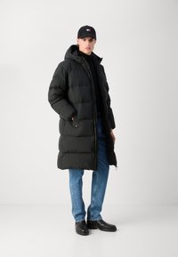 Black padded coat with a hood, long design, featuring side pockets. Worn over a black top, paired with light blue jeans and black boots.