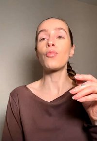 Donna con capelli intrecciati e top marrone che fa una smorfia da bacio, con la mano vicino al viso su uno sfondo semplice.