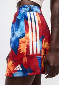 Κλείσιμο εικόνας ενός ατόμου που φοράει πολύχρωμα λουλουδάτα σορτς Adidas με μπλε, κόκκινα και πορτοκαλί λουλούδια και λευκή τριπλή ρίγα της Adidas στο πλάι.