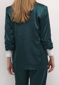 Teal satinblazer med rynkade ärmar, med en bakseam och kontrasterande vita knäppta manschetter. Slät textur, elegant design.