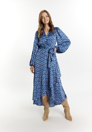 Sinivihreä kuvioitu kietaisumekko, jossa on pitkät hihat, vyötäröä korostava malli ja kerroksellinen hame. Yhdistetty beigeihin kärjellis-ankkleboots-kengiin.