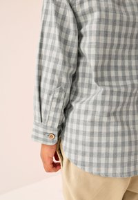 Chemise en coton à carreaux gris et blanc avec un poignet boutonné et un ourlet arrondi, associée à un pantalon beige. La texture semble douce et tissée.