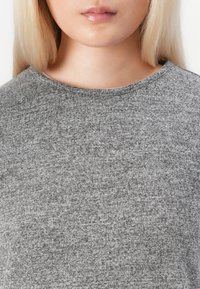 Woman with long blonde hair wearing a grey knit sweater, shown from the neck to upper chest.