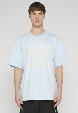 Młody mężczyzna stojący przodem, ubrany w jasnoniebieski, oversize'owy t-shirt z napisem "KARL KANI 89 NEW YORK" oraz czarne plisowane spodnie.