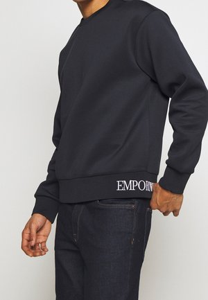 Homme portant un sweat-shirt noir à manches longues avec un logo blanc "EMPORIO" sur l'ourlet, assorti à un jean bleu foncé, posant devant un fond clair uni.