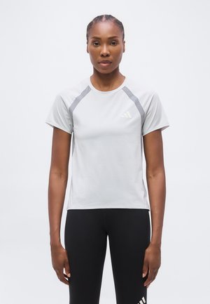 Kvinde med flettet hår iført en lysegrå Adidas kortærmet skjorte og sorte leggings, der står foran en ensfarvet hvid baggrund.