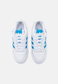White athletic sneakers featuring blue stripe accents, a perforated toe box, and a branded tongue with a floral logo.
