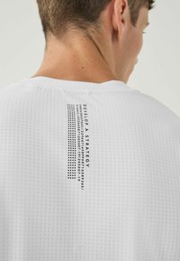 White, textured athletic shirt featuring a printed black graphic with text and numbers on the upper back. Short sleeves and crew neckline.