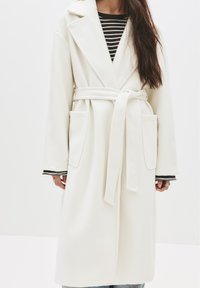 Biela vlnená dlhá bunda s šálovým golierom, predným viazaním a dvomi prednými vreckami. Nosená cez čierno-biele pruhované tričko, ktoré ukazuje rukávy.