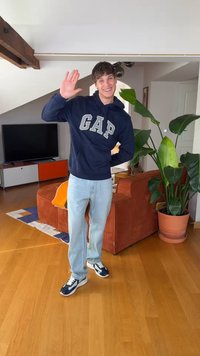 Joven con sudadera azul marino de GAP y jeans claros que saluda sonriente en una sala de estar con un sofá marrón, una planta grande en maceta y un televisor.