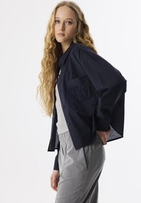 Marineblauw overhemd met knopen en een zak met geborduurde initialen, gedragen over een grijze top en gecombineerd met lichtgrijze broek met elastische tailleband.
