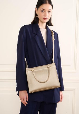 Femme en blazer bleu marine et pantalon portant un sac à main en cuir beige texturé avec bandoulière et double poignée.