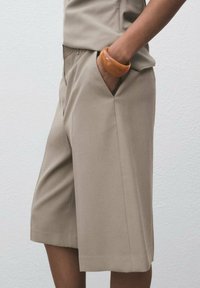 Beige skræddersyede shorts med sidelommer, glat tekstur og en afslappet pasform. Bæret med et rundt, orange-brunt armbånd.