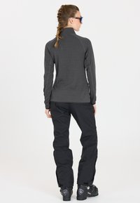 Graues Langarmshirt und schwarze Schneeanzughose mit Reißverschlüssen und Taschen. Schwarze Skistiefel mit Branding. Glatte Stofftextur.