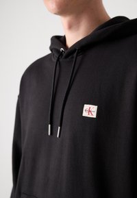 Zwarte hoodie van een katoenmix, met een voorzak, verstelbare trekkoord, zilverkleurige tips op de veters en een klein merklabel.