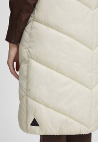 Gilet matelassé crème avec un design en chevron, présentant un logo triangulaire noir en bas et une texture lisse et brillante.