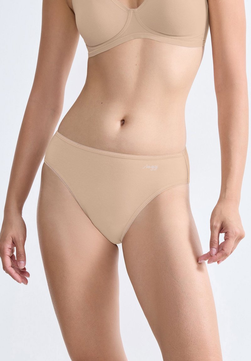 Vrouw in een beige bijpassende beha en hoge slip tegen een effen lichte achtergrond.