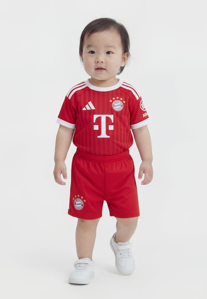 Tout-petit portant le maillot rouge du Bayern Munich avec des baskets blanches, debout sur un fond blanc uni.
